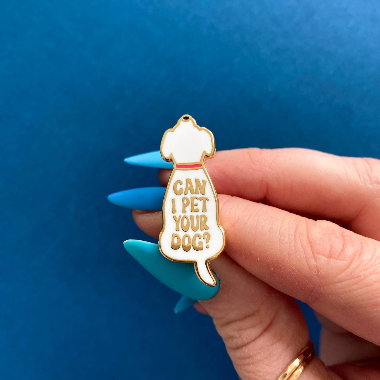 Fingers with stylish blue stiletto nails hold a "Can I Pet Your Dog?" enamel pin against a dark blue background.