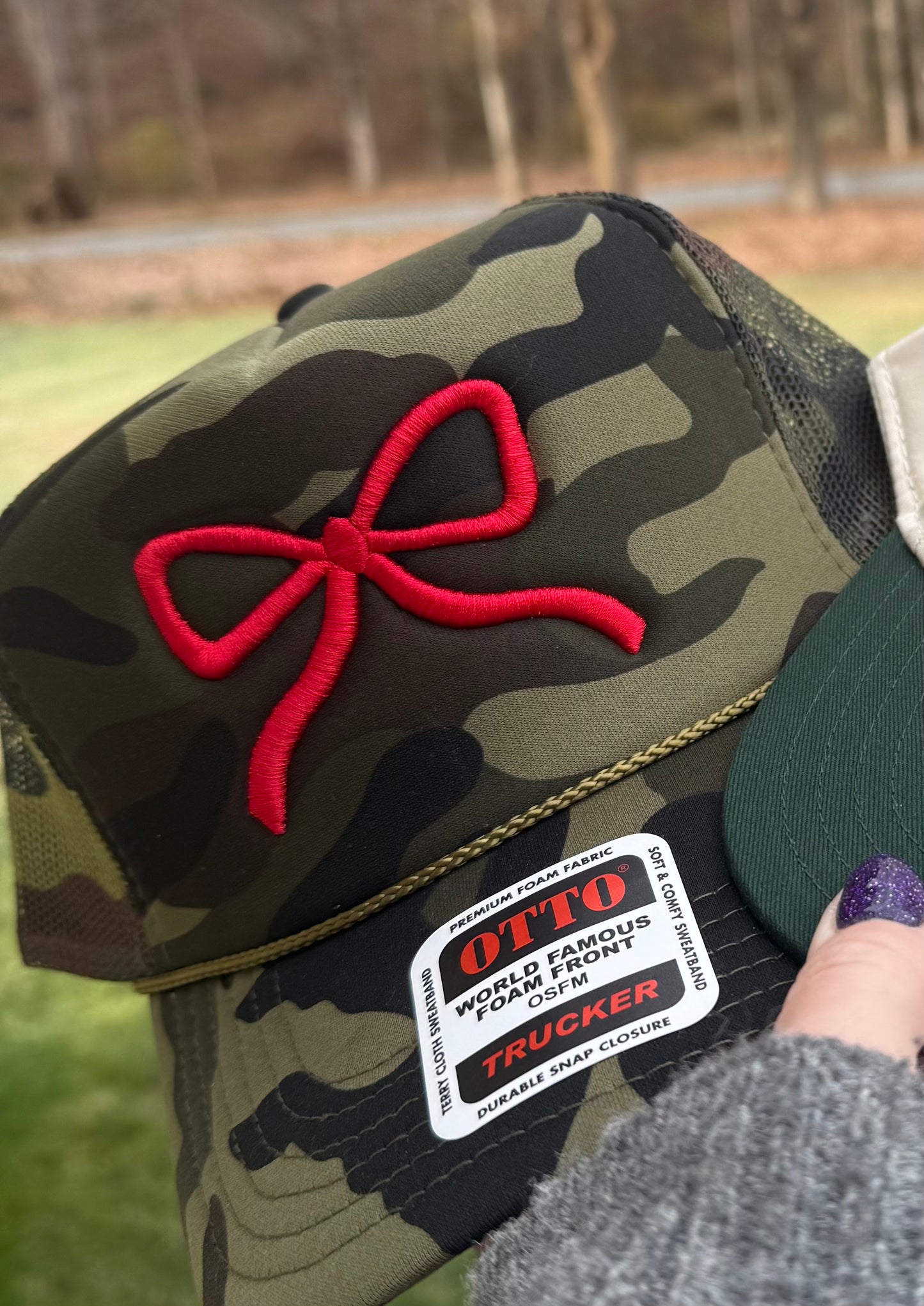 Someone holds an Otto camo trucker hat featuring a red ribbon design outdoors.