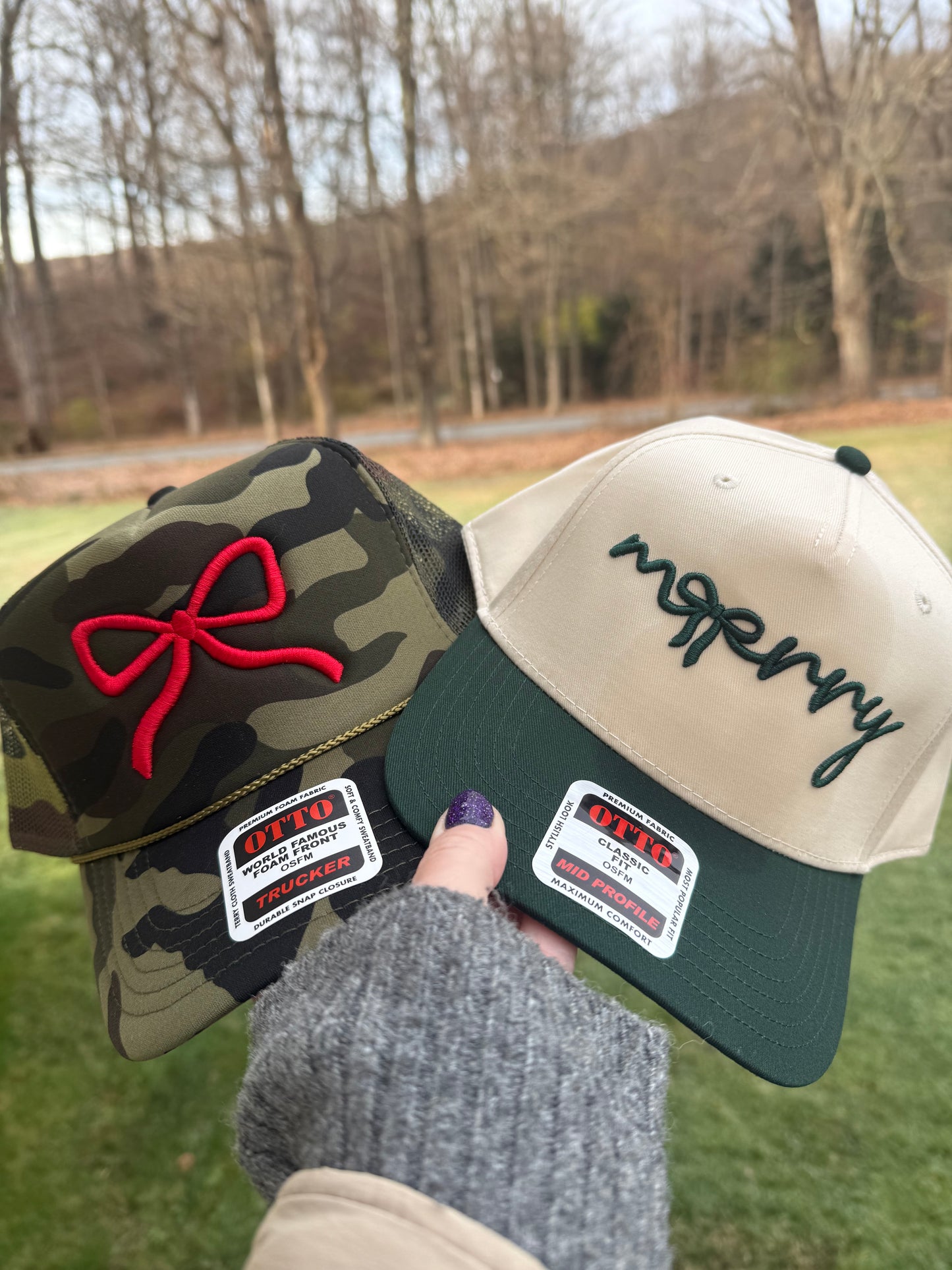 Person holds two Otto brand trucker hats, one camouflage with a red ribbon and one cream with a dark green brim and the word "mopery" embroidered in dark green.